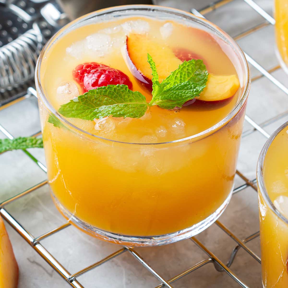 a peach margarita on a countertop with a small shaker, topped with raspberries and other garnishes