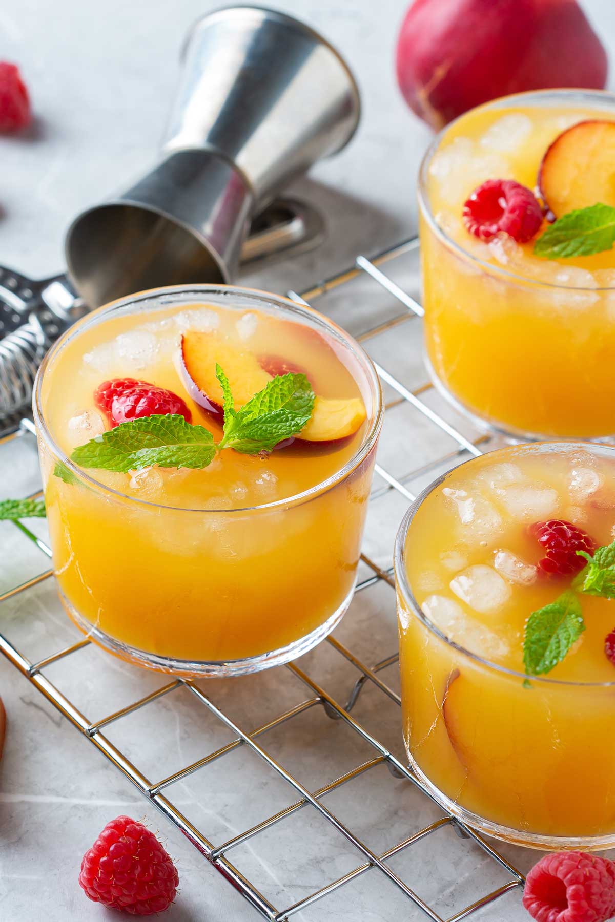 three peach margaritas on a countertop with a small shaker, topped with raspberries and other garnishes