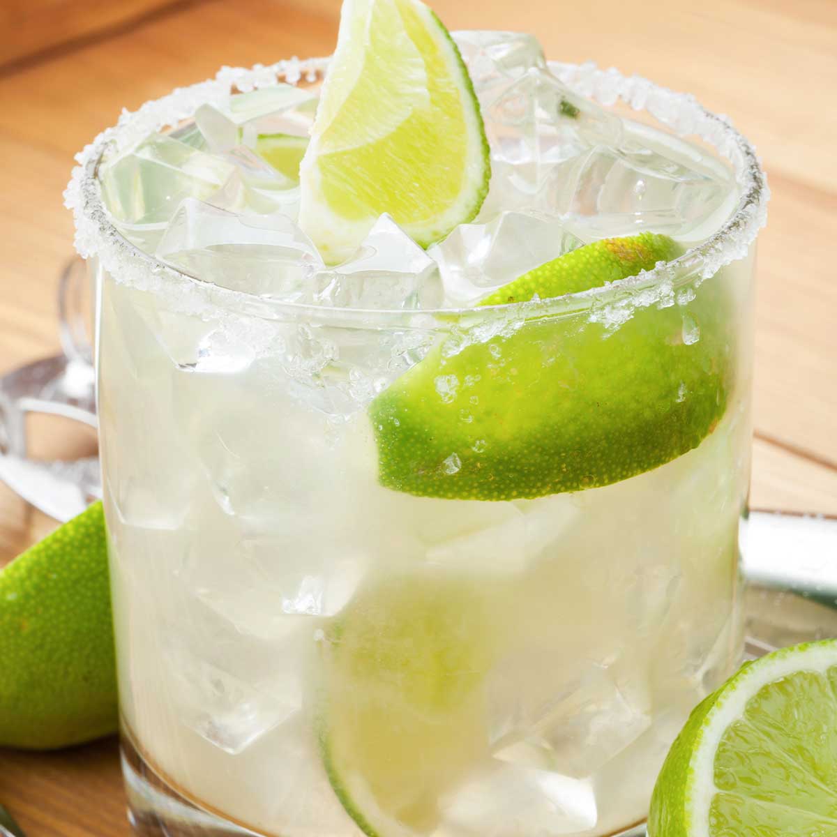 Low-calorie margarita in a salt rimmed glass with wedges of lime, fresh whole lime sliced on wooden countertop.