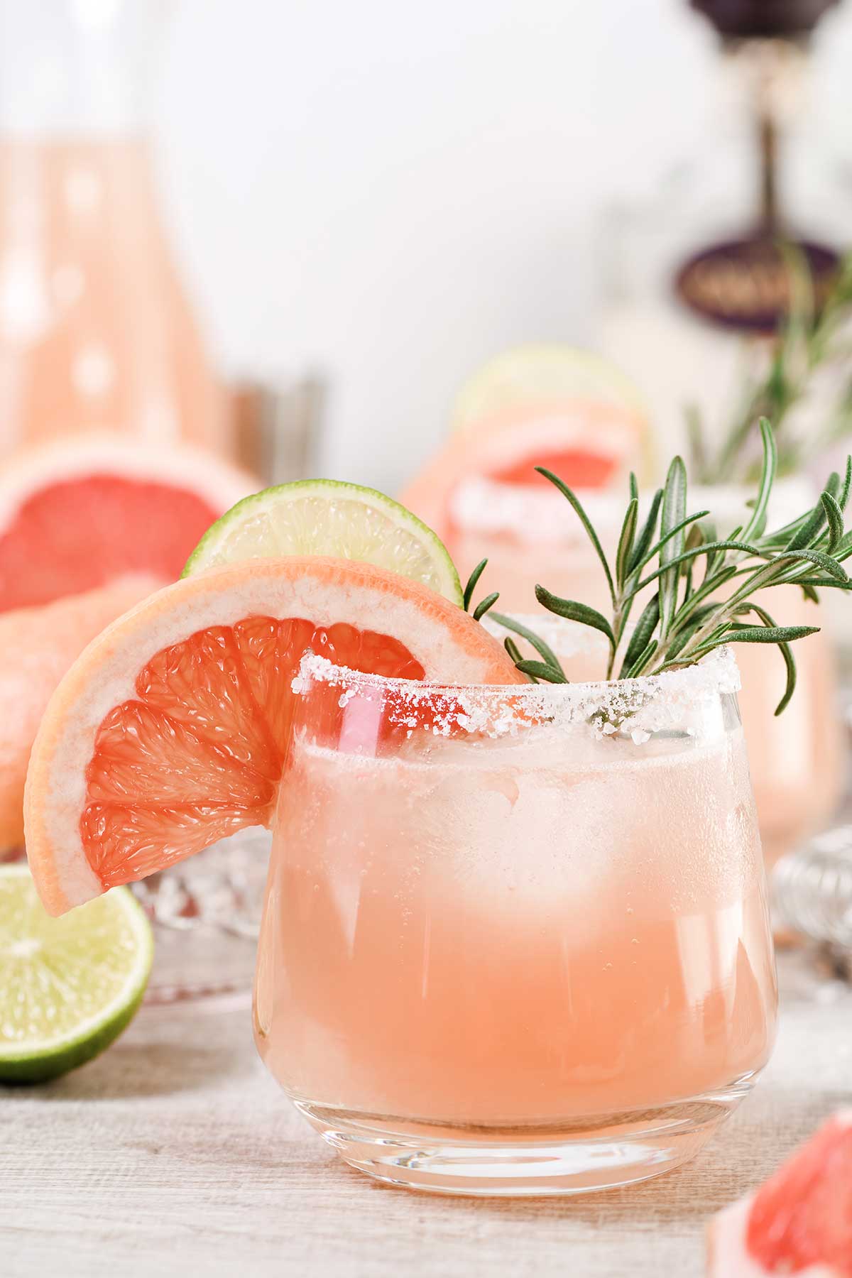 Paloma cocktail in a glass with a salt rim, garnished with a sprig of rosemary, a wedge of grapefruit, and a wedge of lime.