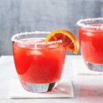 two blood orange margarita glasses with salted rims on a white surface with a wedge of blood orange as garnish