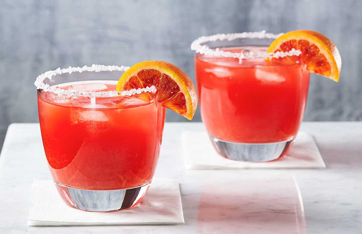 two blood orange margarita glasses with salted rims on a white surface with a wedge of blood orange as garnish