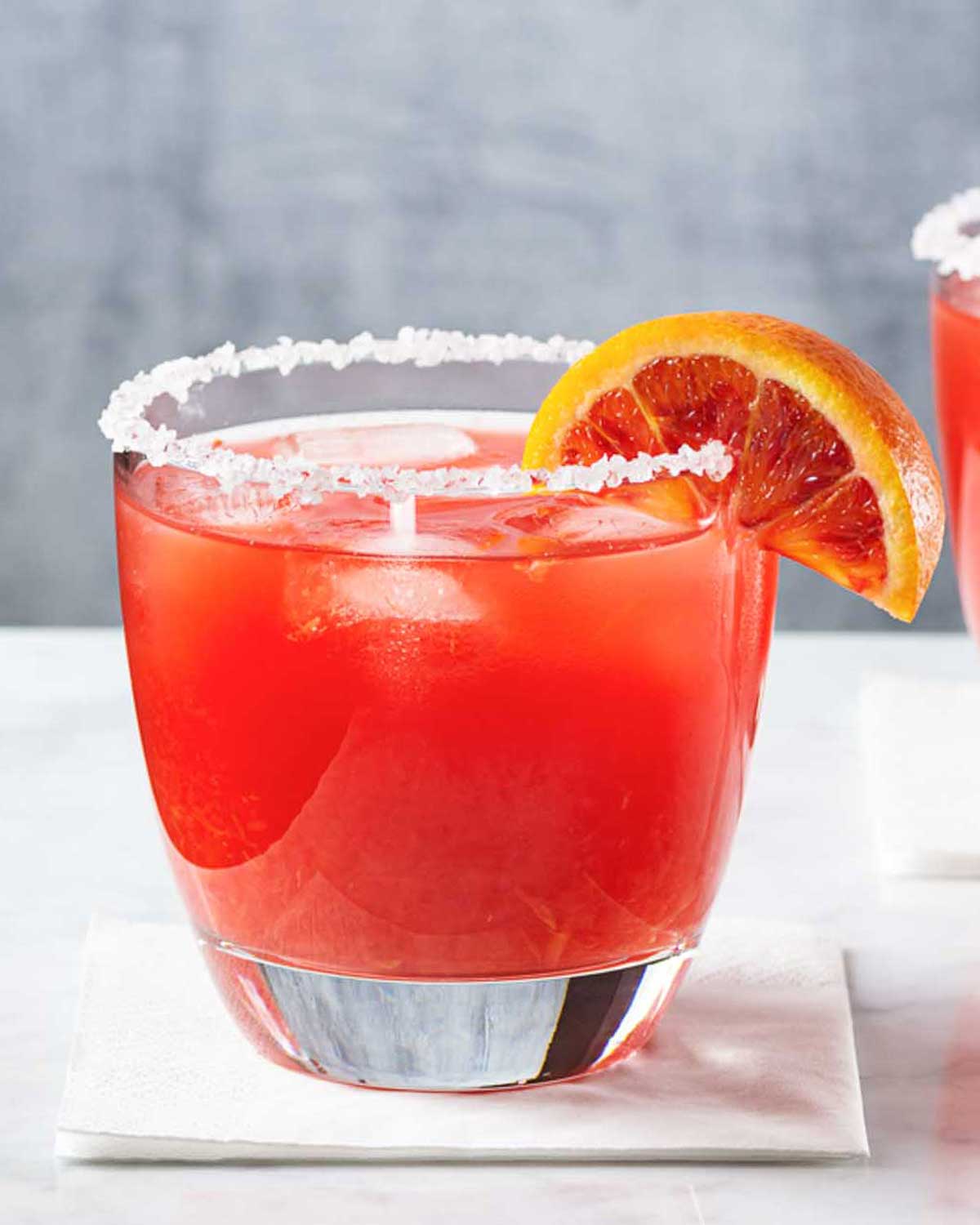 a blood orange margarita glass with salted rim on a white surface with a wedge of blood orange as garnish