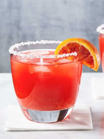 a blood orange margarita glass with salted rim on a white surface with a wedge of blood orange as garnish