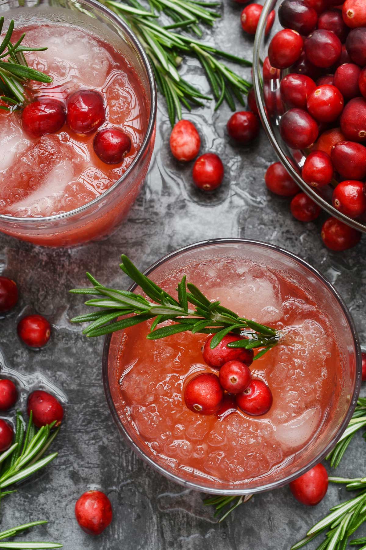 Overhead view of cranberry margaritas with ice, fresh cranberries, and rosemary sprigs, bowl of fresh cranberries.
