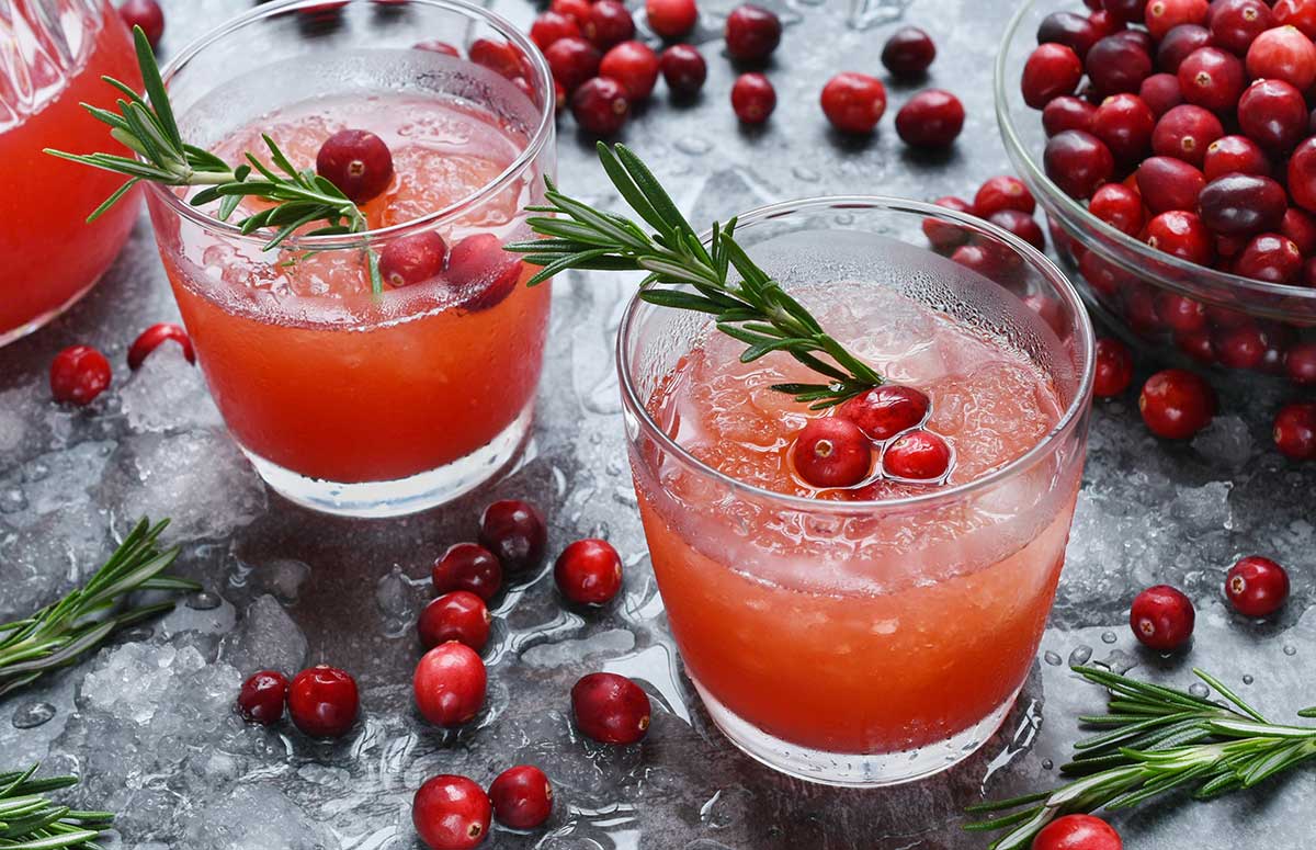Two glasses of cranberry margarita with fresh cranberries and rosemary sprigs, bowl of fresh cranberries, loose cranberries and sprigs on a dark marble countertop.