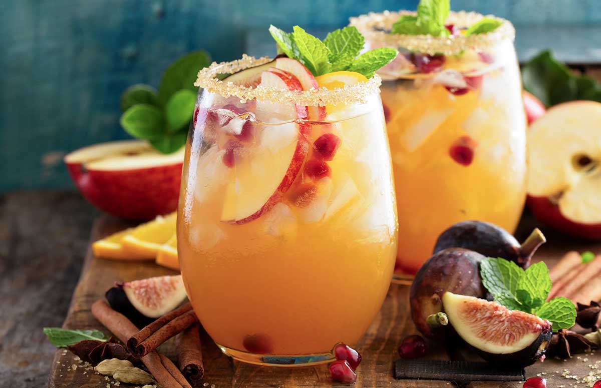 Two cinnamon sugar-rimmed glasses with Fall Sangria Margarita, apple slices, cinnamon sticks, sliced figs, and mint.