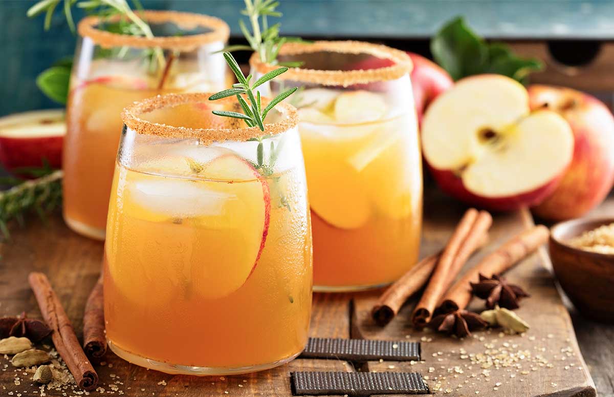Thanksgiving Margarita mixture served in three glasses rimmed with cinnamon sugar and garnished with sliced apple, cinnamon stick, and a sprig of rosemary. Extra ingredients on a wooden kitchen board.