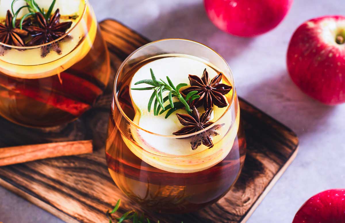Spiced Apple Margaritas with sliced apple, star anise cloves, and a sprig of rosemary, seated on a wooden board.