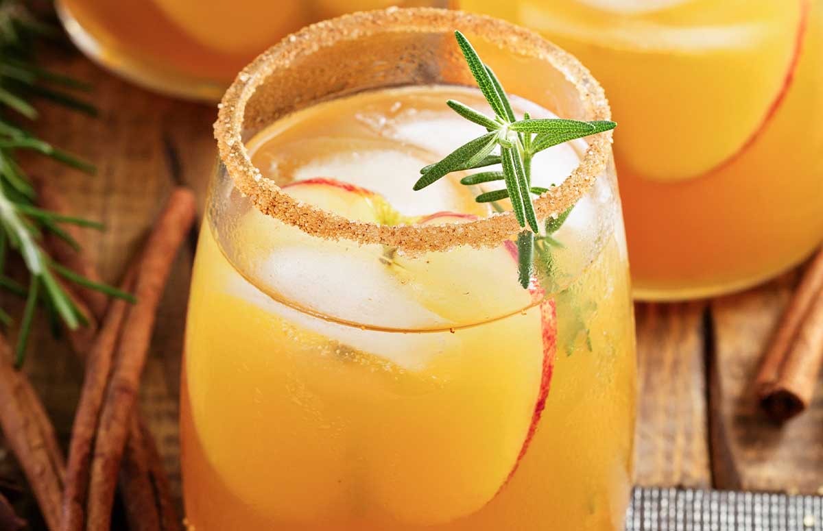 Thanksgiving Margarita mixture served in three glasses rimmed with cinnamon sugar and garnished with sliced apple, cinnamon stick, and a sprig of rosemary.