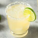 A margarita glass with a salted rim with a margarita mix and a lime wedge on the side