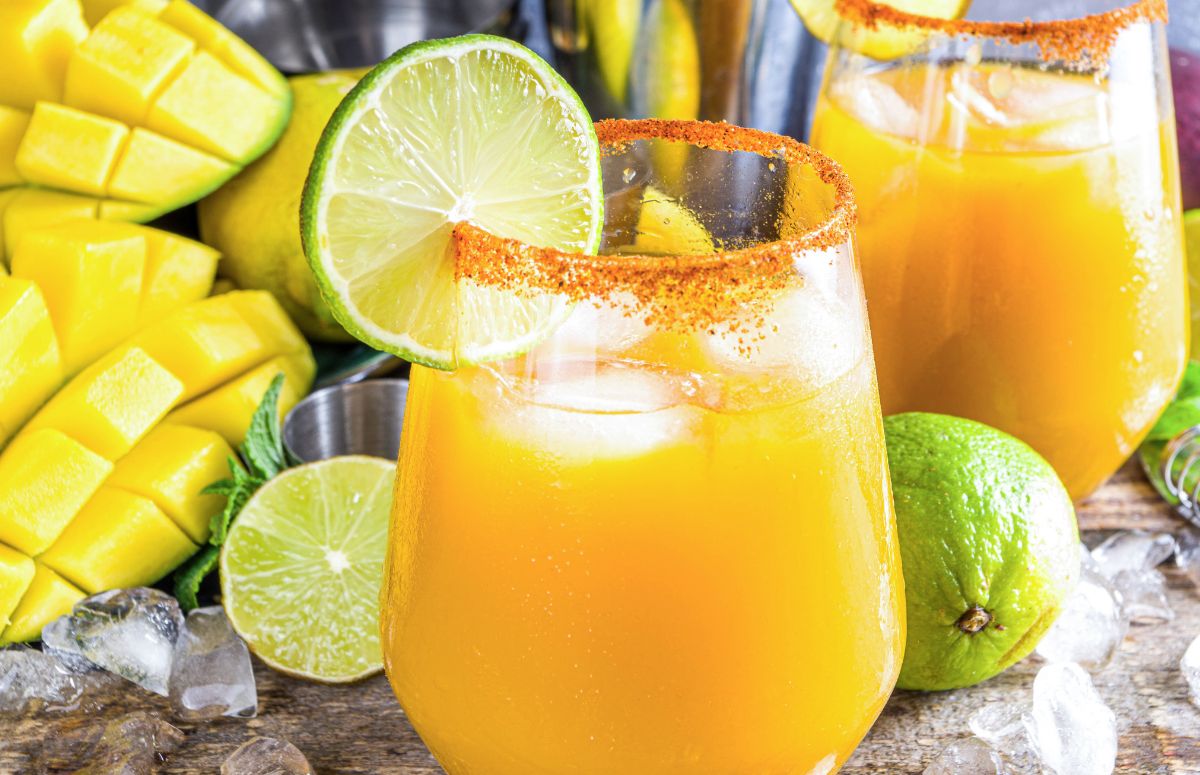 two spicy mango margaritas with tajin rims and lime slice garnish on a countertop with limes and mangoes 